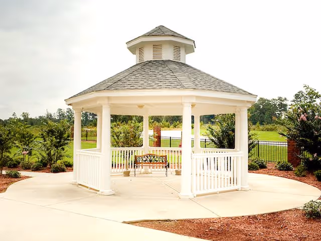 A white gazebo with a shingled roof and open sides situated on a concrete circular patio surrounded by landscaped greenery and trees in an outdoor garden area.