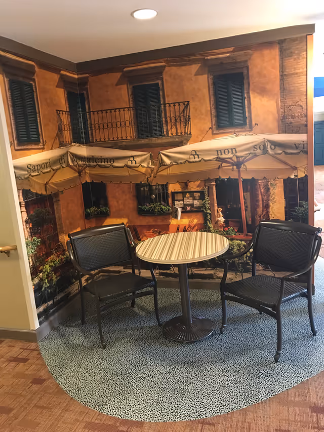 Two black chairs and a round table with a striped top set on a patterned carpet in front of a wall mural depicting a rustic Italian building with balconies and awnings.