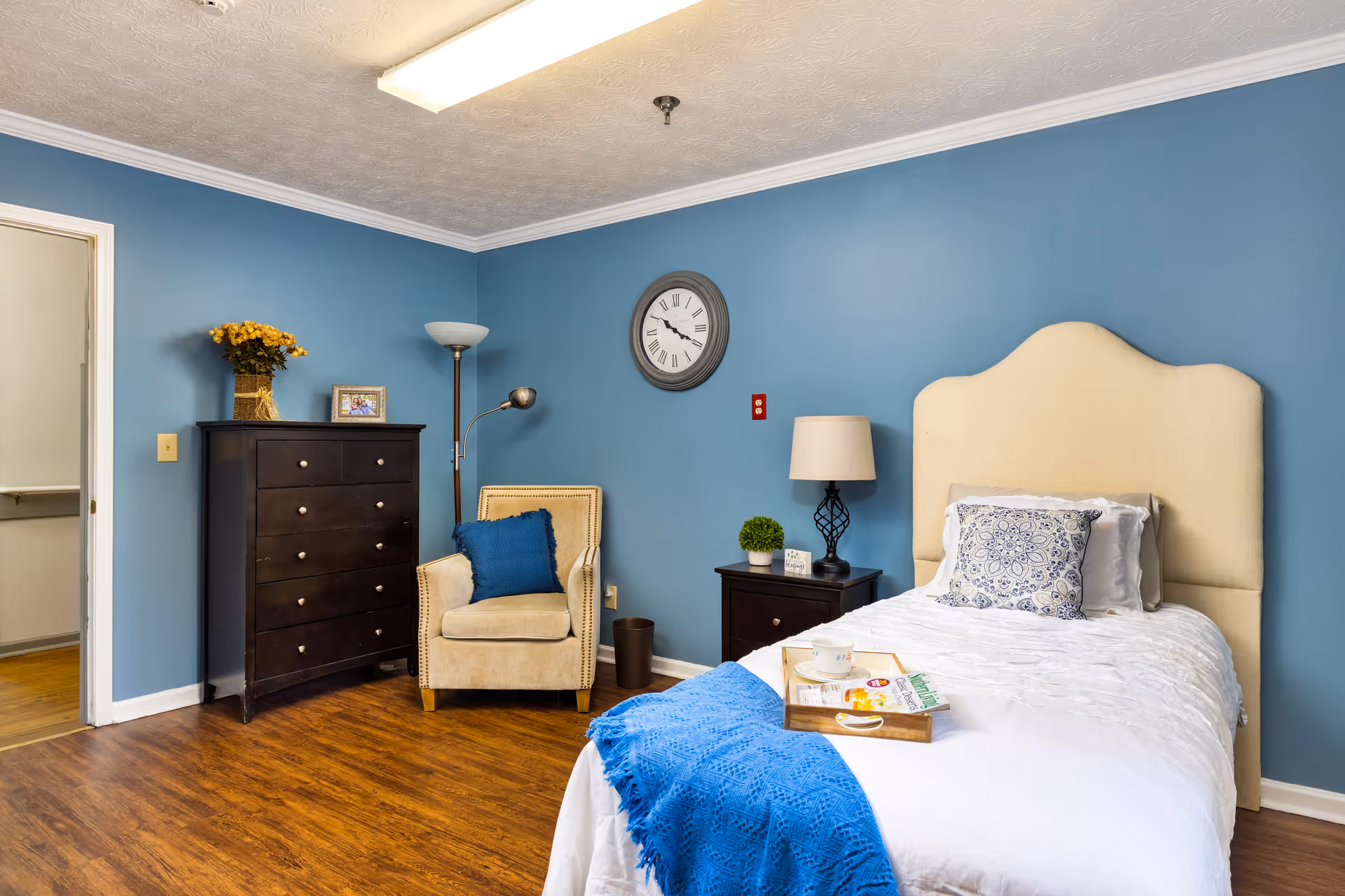 A bright bedroom with a single bed and beige headboard against a blue wall, a dresser, armchair, nightstand, and wall clock.