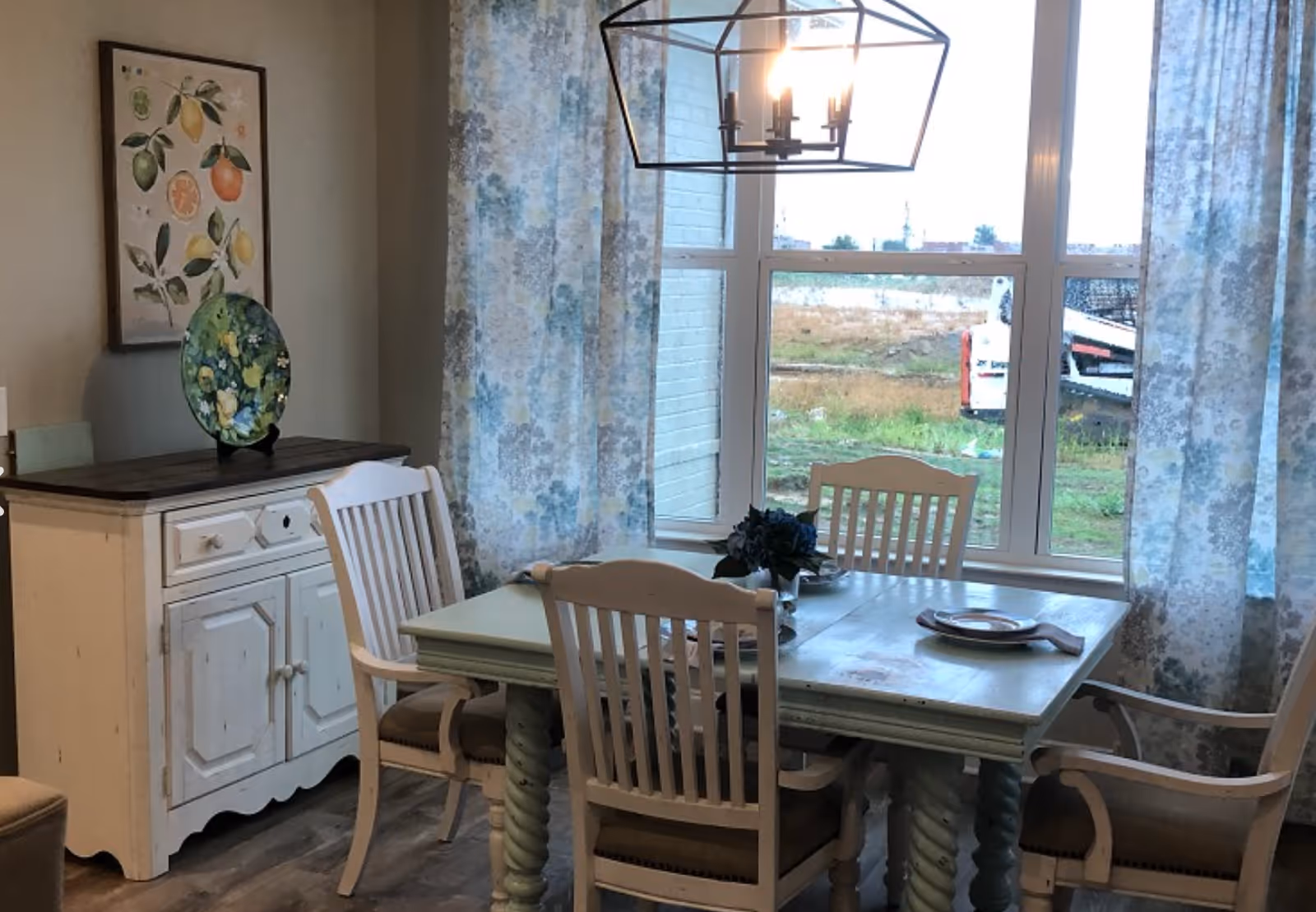 A dining room with a wooden table and four chairs. The table is set with plates and napkins, and there is a small floral centerpiece. Behind the table is a large window with floral curtains, letting in natural light. To the left, there is a white sideboard with a decorative plate and framed botanical artwork above it. A modern chandelier hangs above the table.