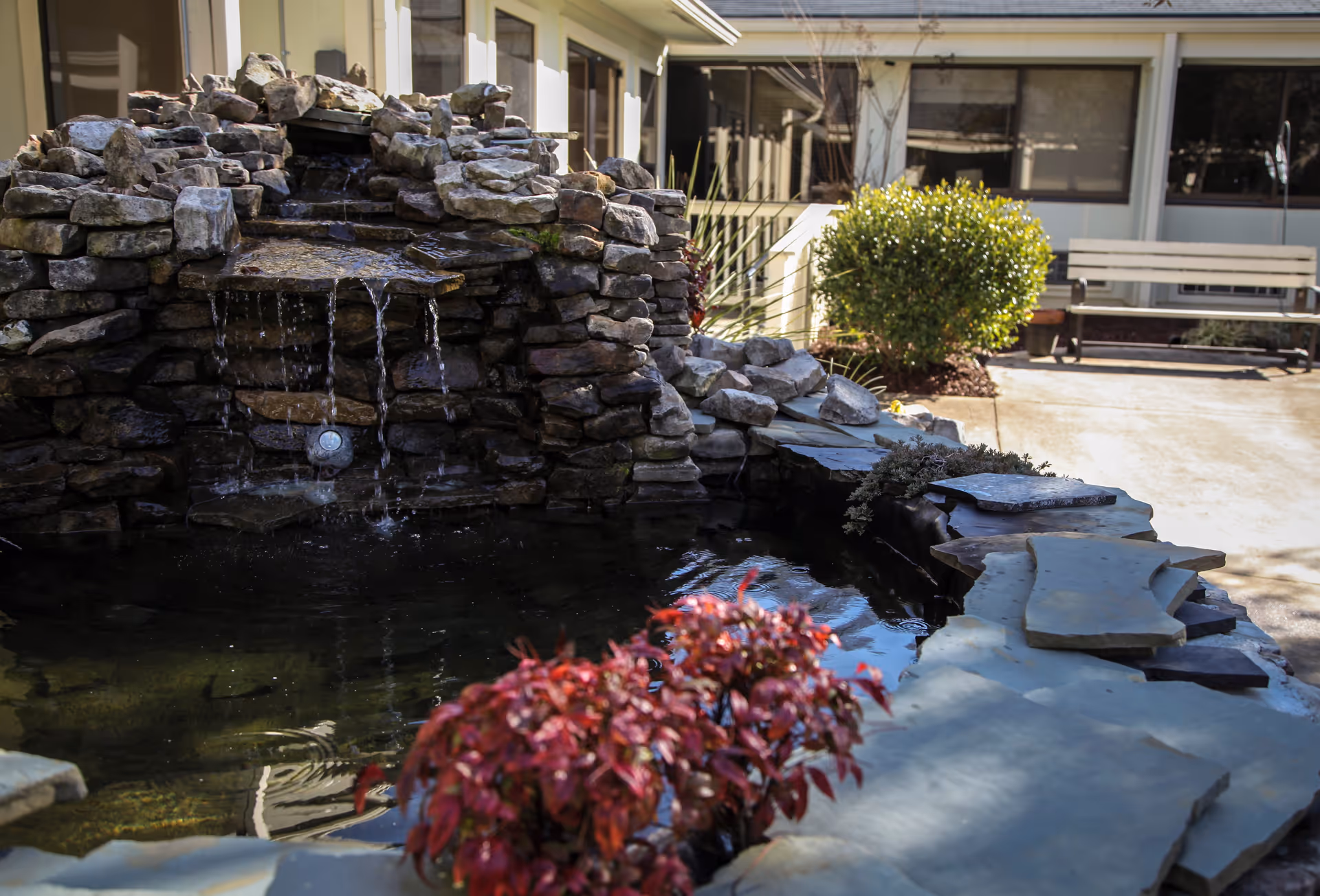 Outdoor stone water feature with a small waterfall flowing into a pond, surrounded by stone edging and plants. In the background, there is a building with windows and a bench on a paved area.