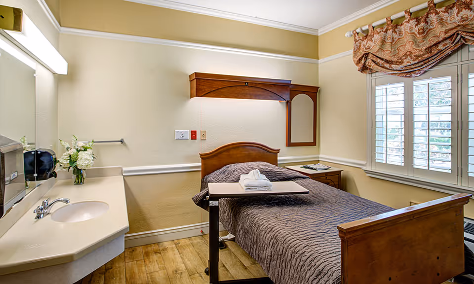 A cozy bedroom in a senior living facility featuring a single wooden bed with a brown quilted blanket, a small overbed table with folded towels, a wooden nightstand with magazines, a wall-mounted wooden cabinet with a mirror, a window with white shutters and patterned valance, and a sink with a mirror and floral arrangement on the left side.
