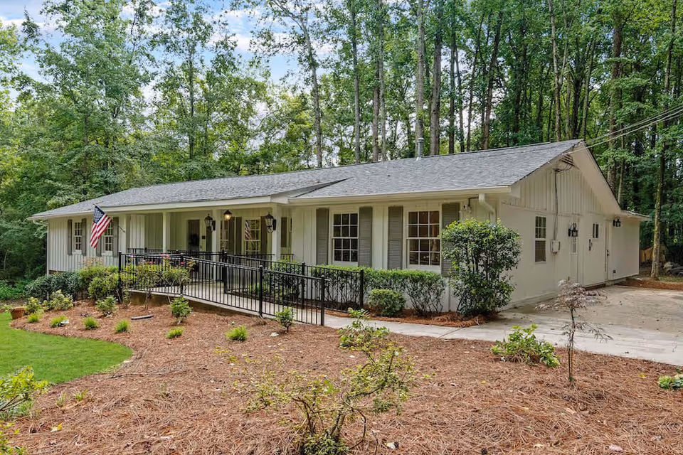 Single-story light-colored ranch-style building with a front porch, wheelchair ramp, American flag, and surrounding trees.