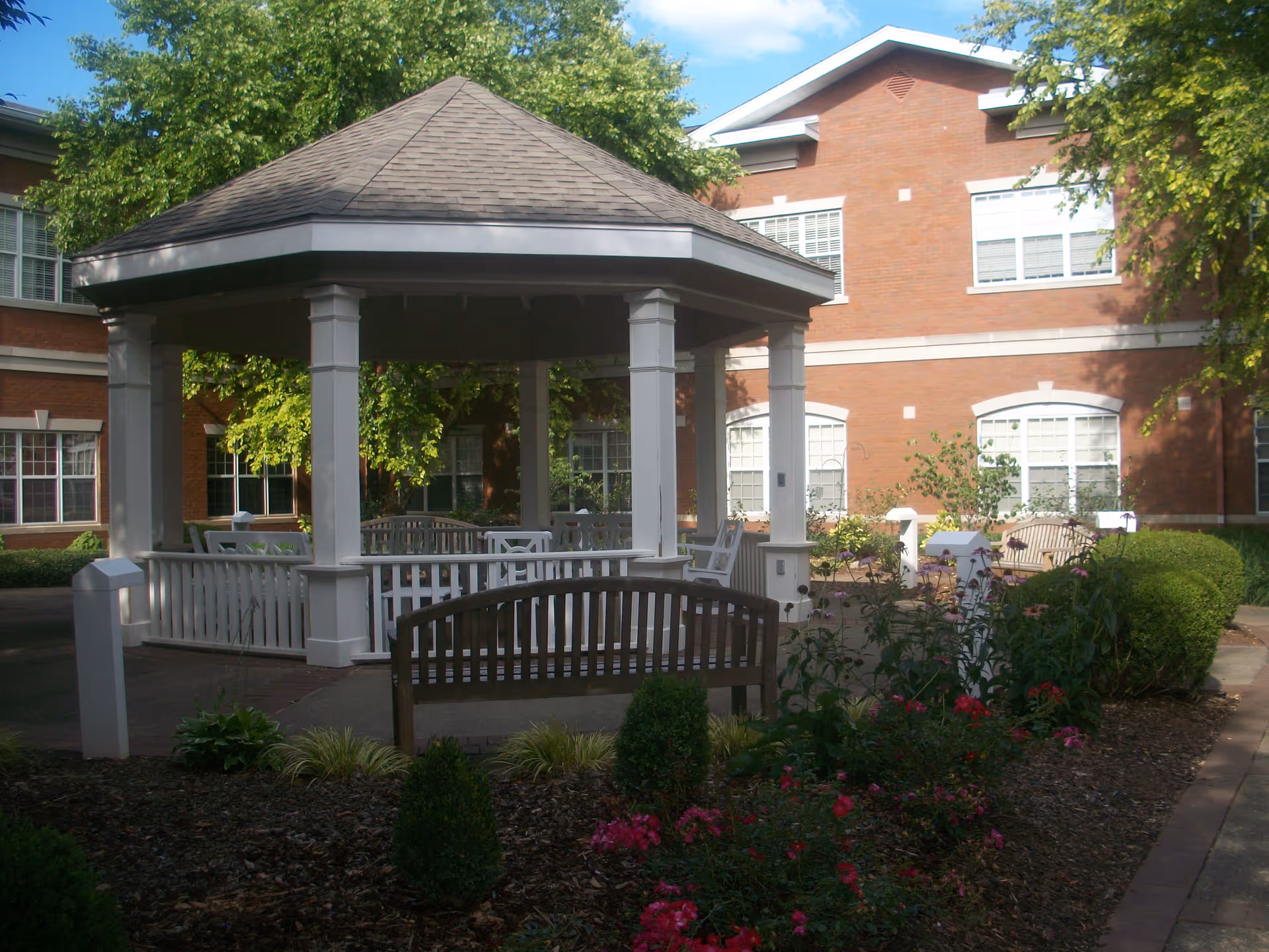 Outdoor courtyard area with a white gazebo surrounded by benches, plants, and flowers. The background shows a red brick building with multiple windows and trees providing shade.