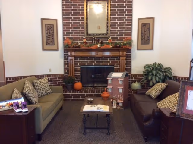 A cozy living room with a brick fireplace in the center, decorated with autumn-themed garlands and pumpkins. Two sofas, one green and one brown, face each other with patterned cushions. A small coffee table with an open book sits between the sofas. There are framed artworks on the walls and a large plant next to the brown sofa.