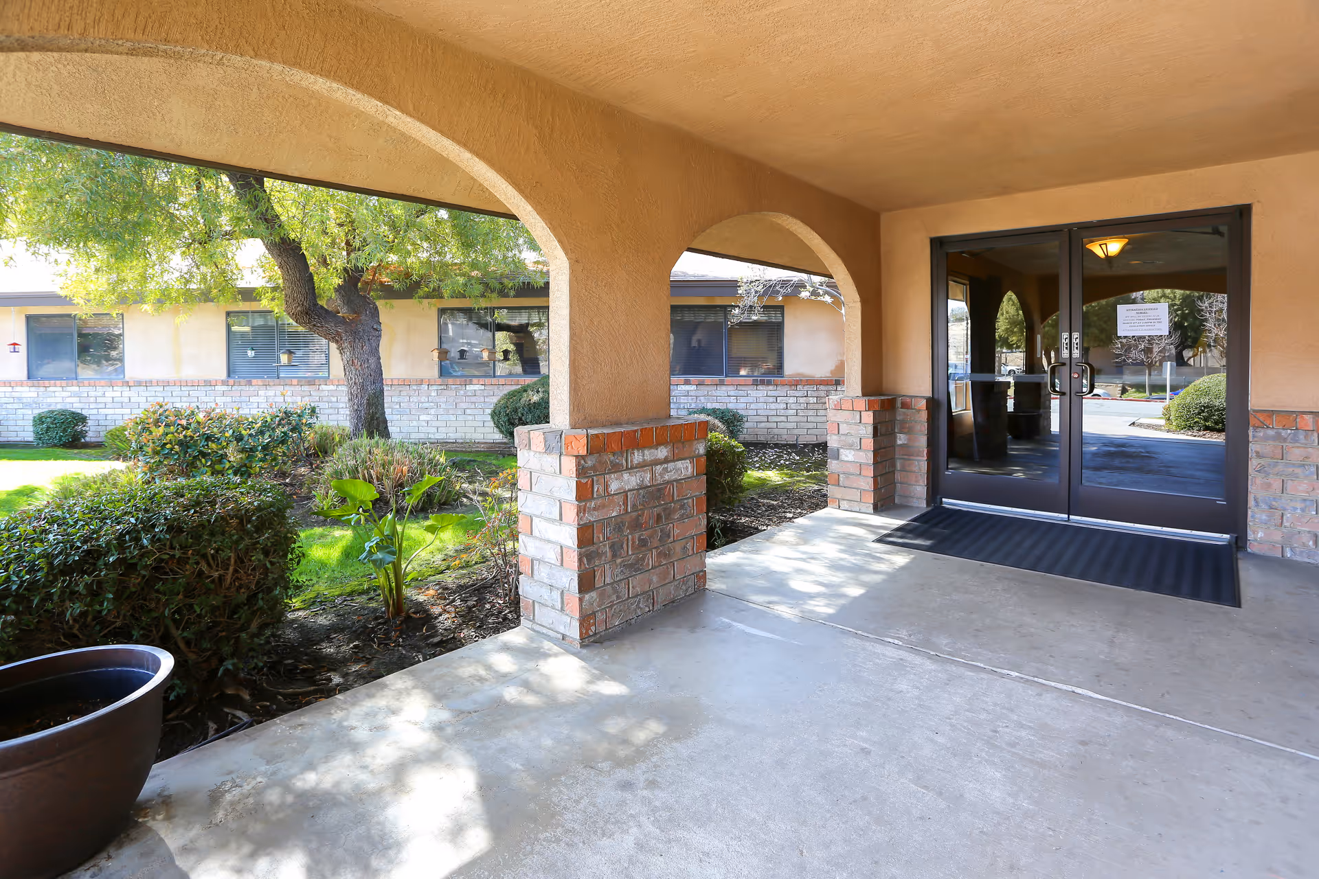 Covered entrance area with brick pillars and arches leading to glass double doors of a building. There are bushes, a tree, and a grassy area visible outside under natural daylight.