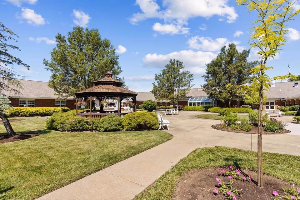 Outdoor courtyard area at Sycamore Run Nursing & Rehabilitation Center featuring a wooden gazebo surrounded by green bushes, trees, and well-maintained grass. There are concrete walkways, white benches, and a clear blue sky with some clouds.