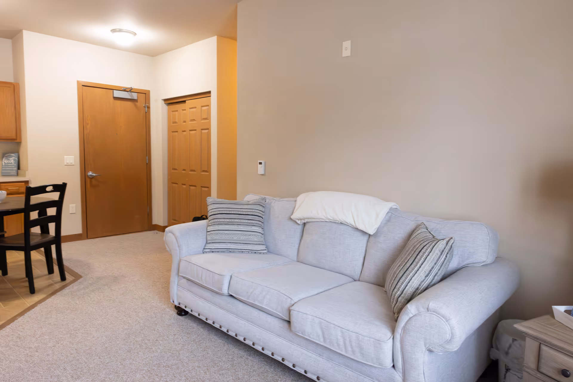 A cozy living room area featuring a light gray upholstered sofa with three cushions and two striped throw pillows. A white blanket is draped over the back of the sofa. To the left, there is a small dining area with a black table and chairs, and wooden cabinets. The walls are painted beige, and there are two wooden doors in the background, one solid and one closet-style with panels. The floor is carpeted in a light color.