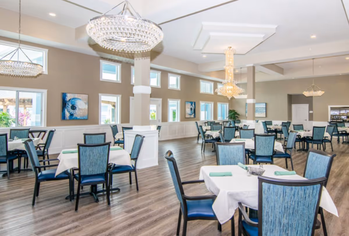 A spacious and well-lit dining room with multiple tables covered in white tablecloths, each set with green napkins and chairs upholstered in blue fabric. The room features large windows, modern chandeliers, light-colored walls, and wood flooring.