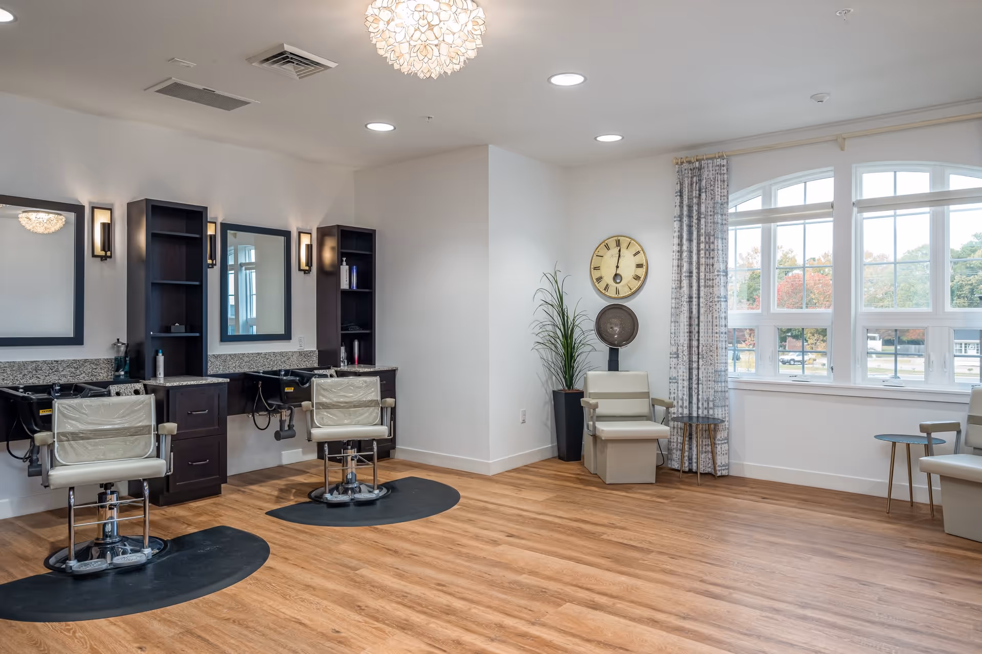 A bright and spacious salon area in a senior living facility with two styling chairs in front of mirrors and sinks, a hair dryer chair, a large wall clock, a tall plant, and large windows with patterned curtains letting in natural light.