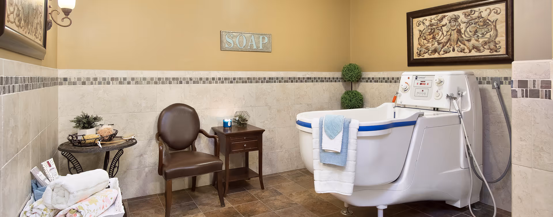 A well-lit bathroom with beige tiled walls and floor. There is a white walk-in bathtub with blue trim on the right side, with white and blue towels hanging over the edge. Next to the tub is a small wooden side table with a lit candle and a decorative glass container. A brown leather chair is positioned beside the table. On the left side, there is a small round table with a basket containing bath items and a potted plant. Rolled towels and bath linens are stacked nearby. The walls are decorated with framed artwork and a sign that reads 'SOAP'.