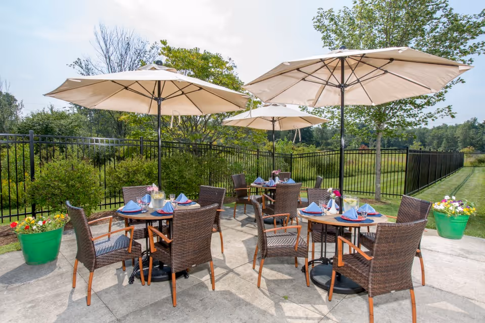 Outdoor patio area with several round tables set for dining, each with four wicker chairs and large beige umbrellas providing shade. The tables have blue napkins, glasses, and pitchers of water with lemon slices. The patio is surrounded by greenery, including bushes, trees, and potted flowers, with a black metal fence enclosing the area.