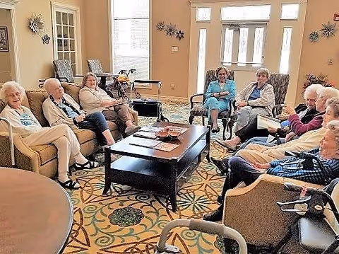 A group of elderly people sitting in a living room area with armchairs and sofas arranged around a central coffee table. The room has large windows and a door with glass panels letting in natural light. The floor is covered with a patterned carpet, and there are decorative wall hangings.