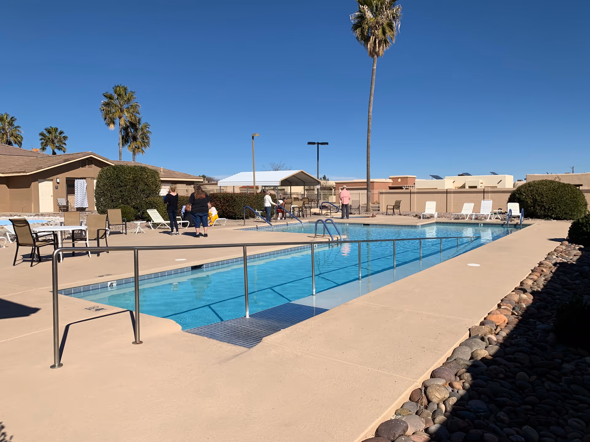 Outdoor swimming pool area at Vista View Resort with several people standing and sitting near the pool. There are lounge chairs, tables with chairs, palm trees, and a clear blue sky in the background.