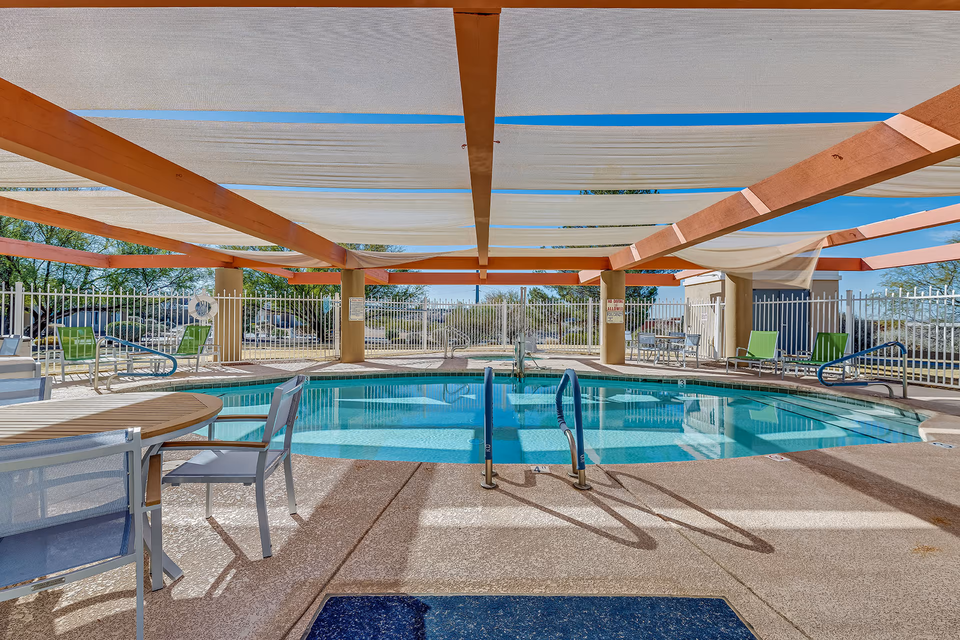 Outdoor swimming pool area at Solstice Senior Living at Las Cruces with a shaded pergola overhead, pool handrails, tables and chairs around the pool, and a fenced perimeter with desert landscaping visible beyond.