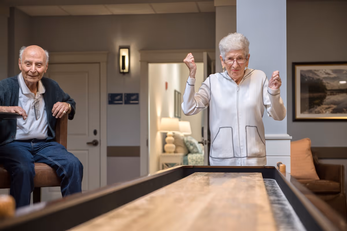 Two elderly people play shuffleboard in a well-lit common area of a senior living facility.