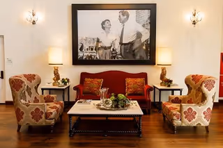 A cozy living room area with a red upholstered sofa and two patterned armchairs facing a rectangular coffee table. The coffee table has decorative items including a small plant and a glass pitcher. Two side tables with matching lamps flank the sofa. A large black and white framed photograph of a man and woman hangs on the wall above the sofa. The room has wooden flooring and wall sconces with lights on either side of the photograph.