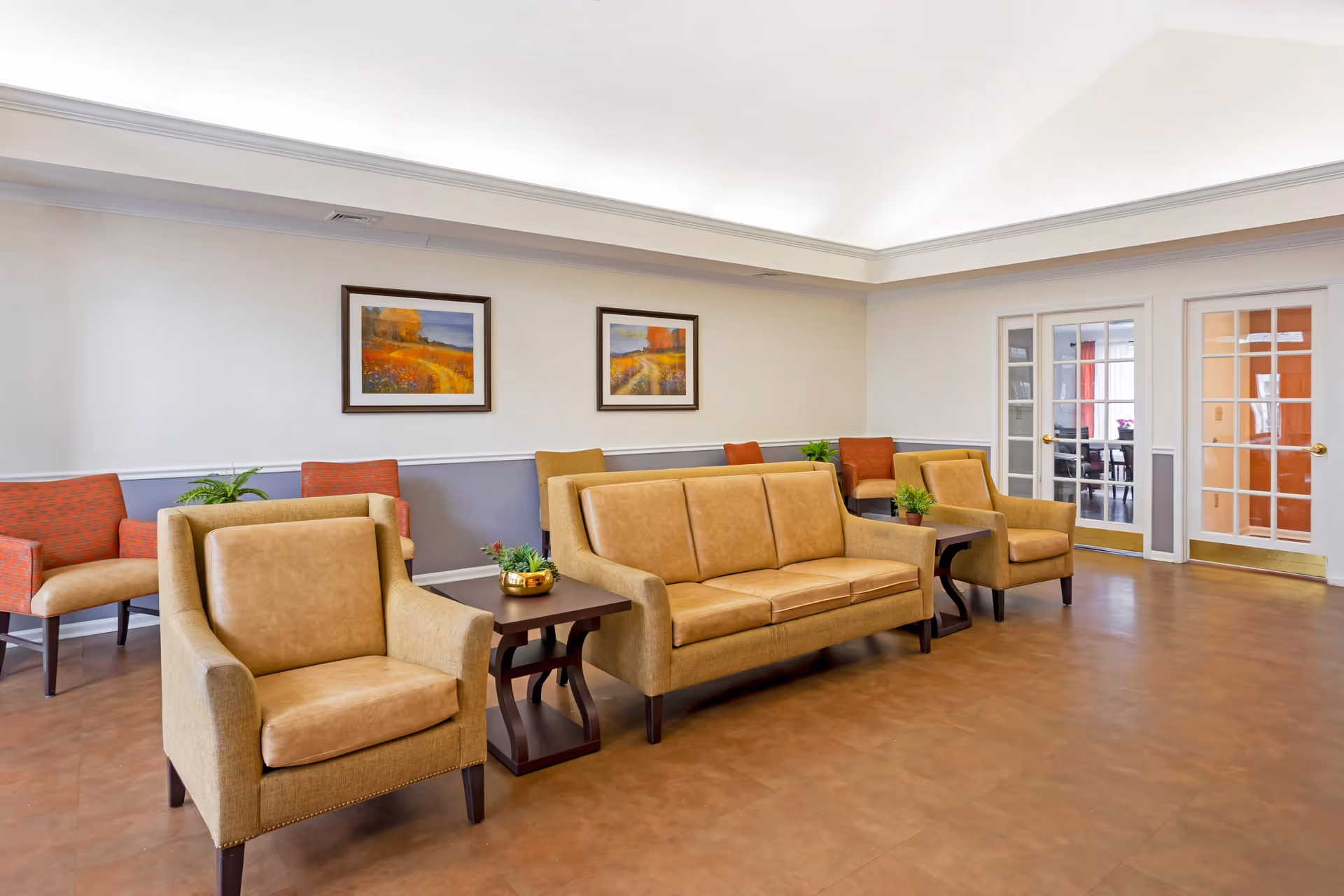 A well-lit living room area with tan leather armchairs and a matching sofa arranged around dark wooden side tables with small potted plants. The walls are painted white with a gray lower half and decorated with two framed landscape paintings. In the background, there are glass-paneled double doors leading to another room.