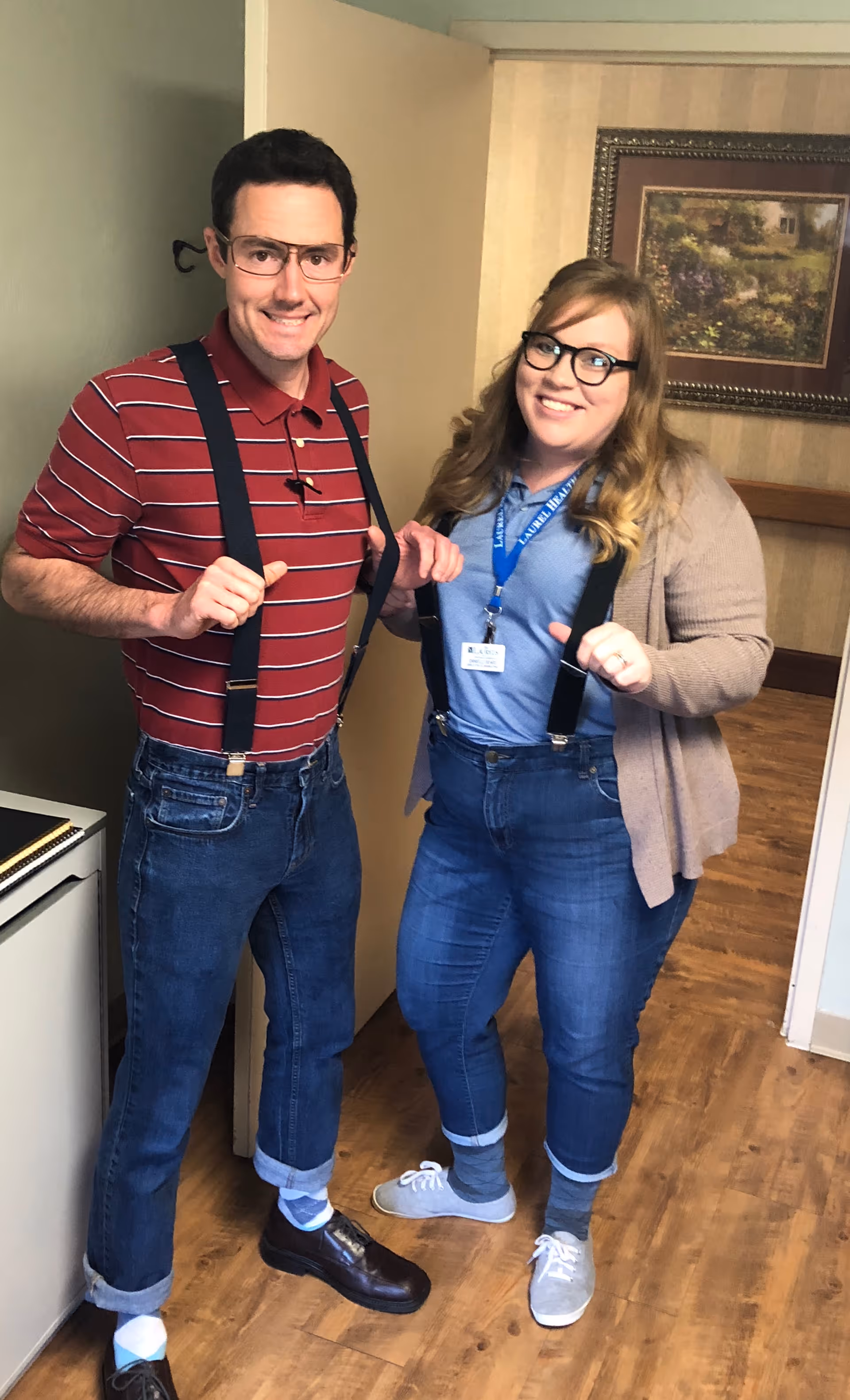 Two people standing indoors on a wooden floor, both wearing glasses, suspenders, rolled-up jeans, and socks with shoes. One person is wearing a red striped polo shirt and dark shoes, while the other is wearing a blue shirt, beige cardigan, and gray sneakers. There is a framed painting on the wall behind them.