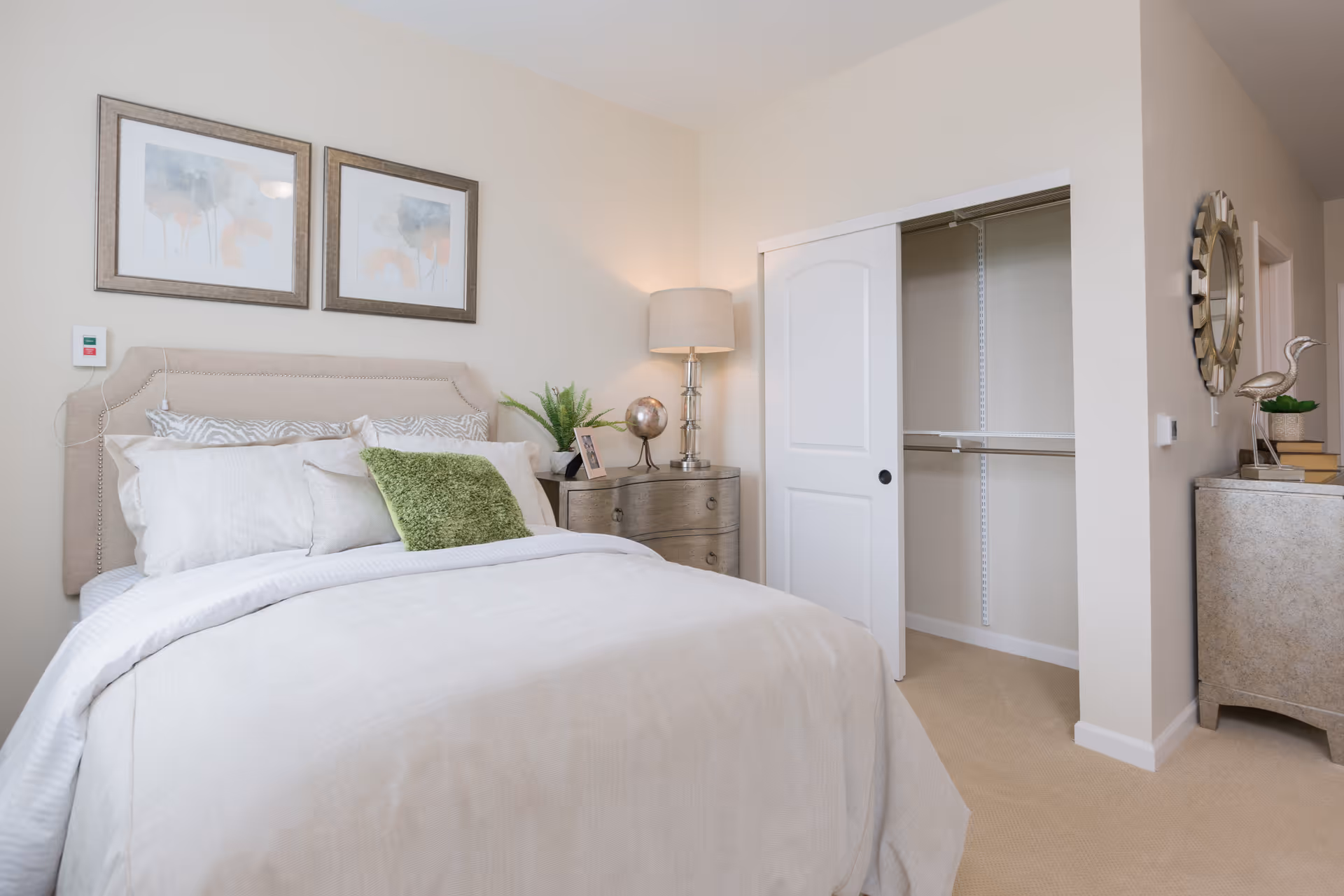 A neatly made bed with white and beige bedding and a green accent pillow in a bedroom. Above the bed are two framed abstract paintings. To the right of the bed is a wooden nightstand with a lamp, a small globe, a framed photo, and a plant. An open closet with sliding white doors is visible, along with a decorative mirror and a small cabinet with books and a bird sculpture on top.