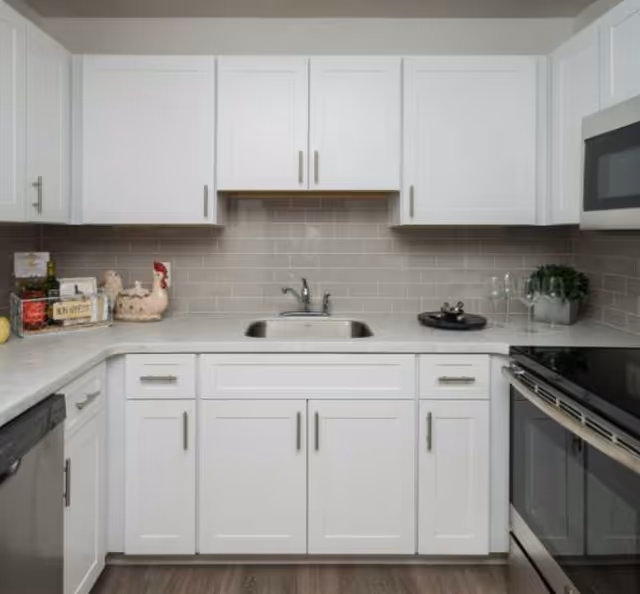 A modern kitchen with white cabinets and drawers, a stainless steel sink with a faucet, a gray tiled backsplash, and a light-colored countertop. On the left side, there are kitchen items including a decorative chicken, bottles, and a basket. On the right side, there is a black stove with an oven and a microwave above it, along with two wine glasses and a small potted plant.