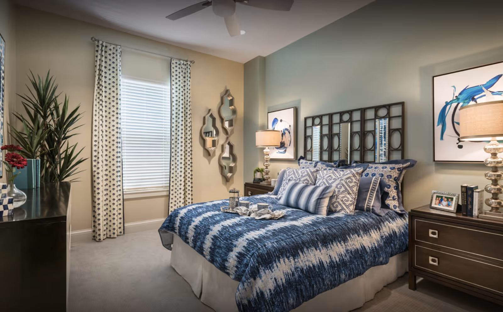 A neatly made bed with blue and white patterned bedding and multiple decorative pillows in a bedroom. The room features a window with white blinds and patterned curtains, a dark wooden dresser with a plant and decorative items, two matching nightstands with lamps, framed artwork on the walls, and three decorative mirrors on one wall.
