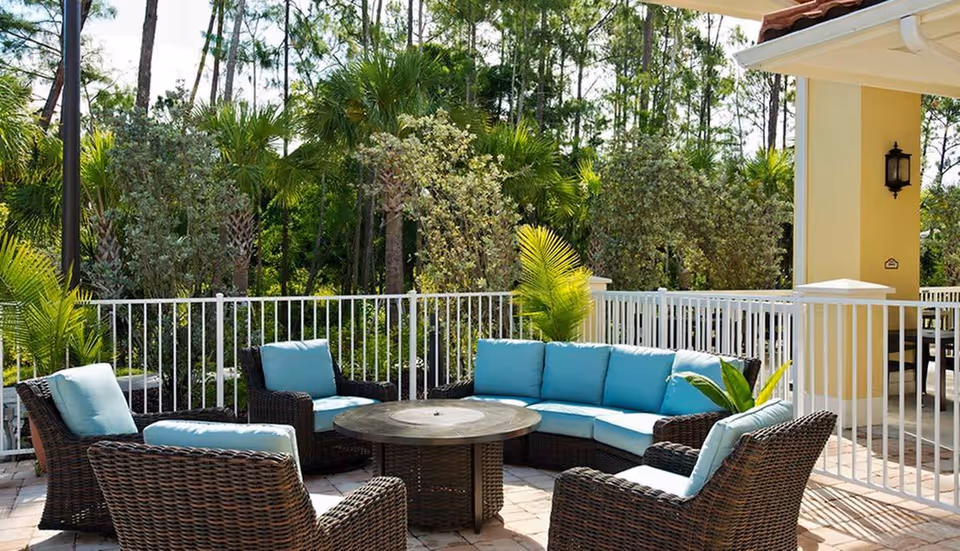 Outdoor patio area with wicker furniture including a curved sofa and several chairs with light blue cushions, a round central table, surrounded by a white metal fence and lush green trees and plants in the background.