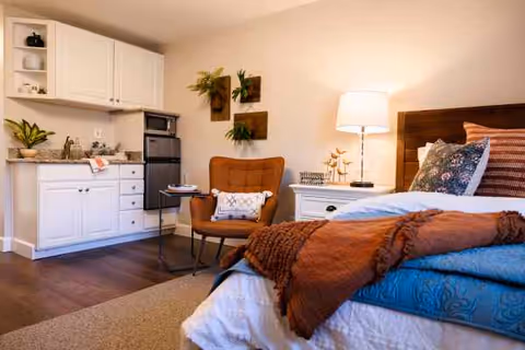 A cozy senior living studio apartment featuring a bed with colorful pillows and a rust-colored throw blanket, a white nightstand with a lamp and decorative items, a comfortable brown armchair with a pillow, and a small kitchenette with white cabinets, a sink, a microwave, and a mini fridge. The walls have decorative plant art, and the flooring is a combination of carpet and wood.