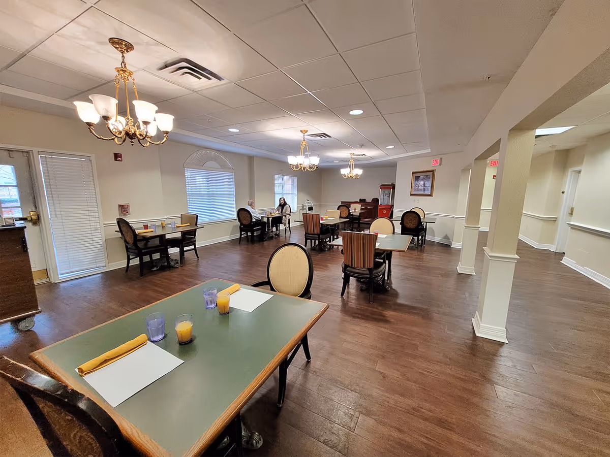 A spacious dining room with multiple tables and chairs arranged neatly. The tables have place settings including napkins, glasses, and placemats. The room features wooden flooring, cream-colored walls, large windows with blinds, and elegant chandeliers hanging from the ceiling. Two people are seated at a table in the background.