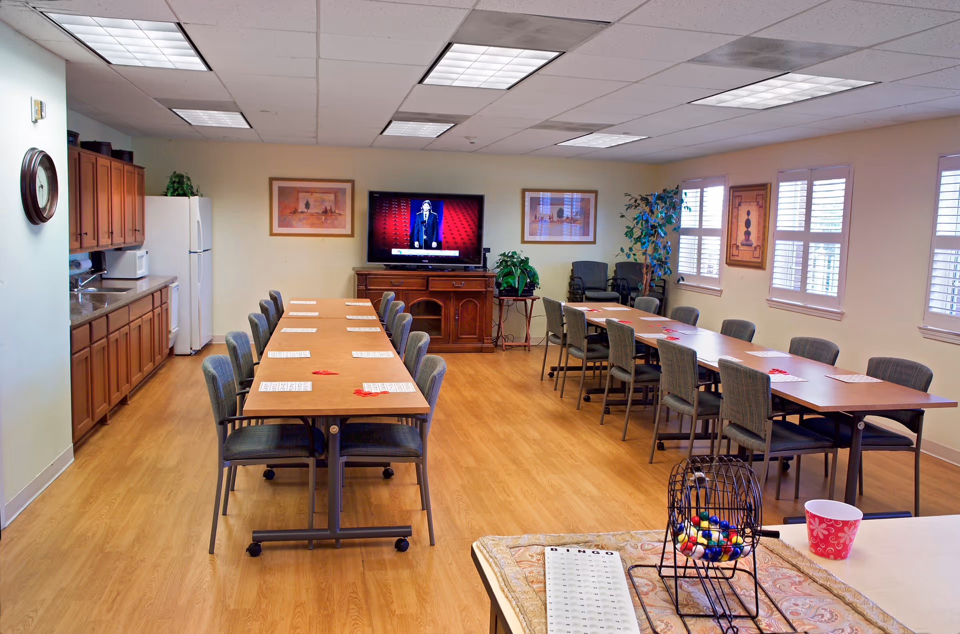A spacious activity room with two long tables arranged in a U-shape, surrounded by chairs. On the tables are bingo cards and red markers. A bingo cage with colorful balls and a bingo card are visible in the foreground. The room has wooden flooring, a kitchenette with cabinets, a refrigerator, and a microwave on the left side. A flat-screen TV is mounted on the far wall, flanked by framed artwork and plants. Windows with white shutters allow natural light into the room.