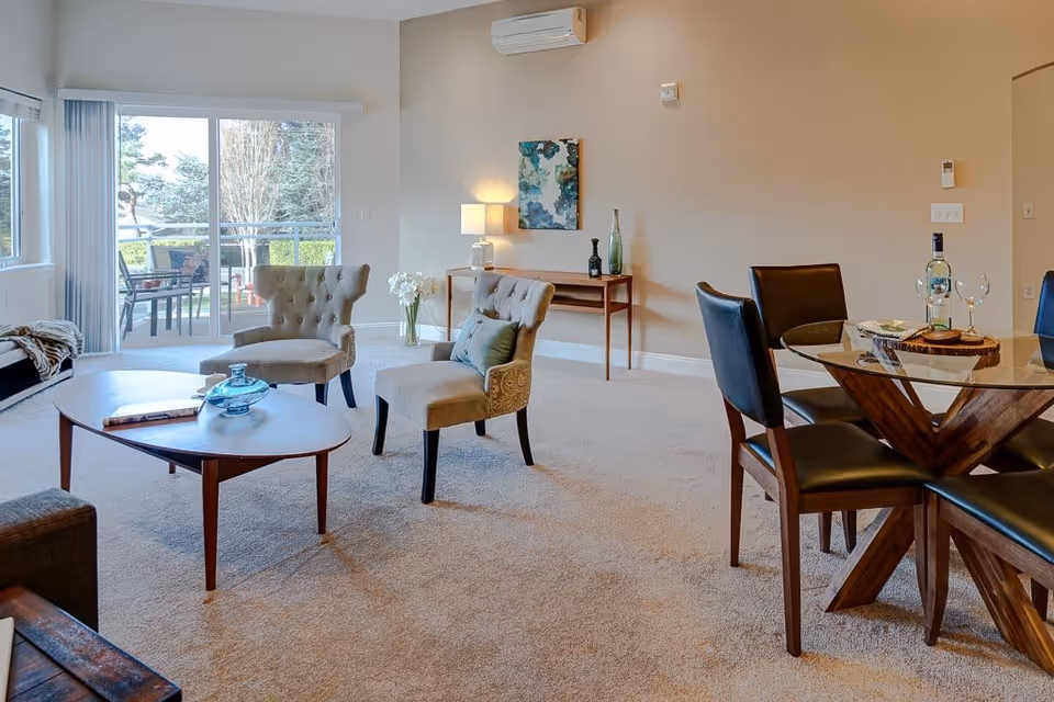 A bright and spacious living room area with beige carpet, two upholstered chairs, a wooden coffee table with a blue glass vase and a book, and a glass dining table with four black leather chairs. There is a sliding glass door leading to an outdoor patio with chairs and greenery visible outside. A small wooden console table with decorative items and a lamp is against the wall, along with a piece of abstract artwork above it.