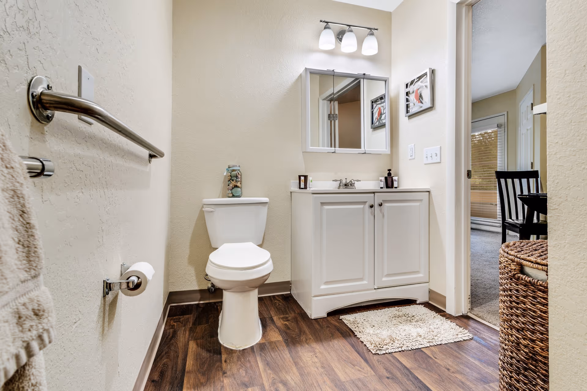 Small bathroom with a toilet, white vanity and mirror, towel bar, and wood-look flooring with an open doorway to a dining area.