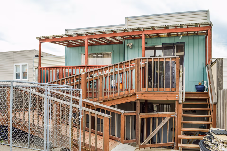 Exterior view of a two-story building with a wooden ramp and stairs leading to a covered porch area. The building has teal siding and a sliding glass door on the upper level. A chain-link fence is visible in the foreground.