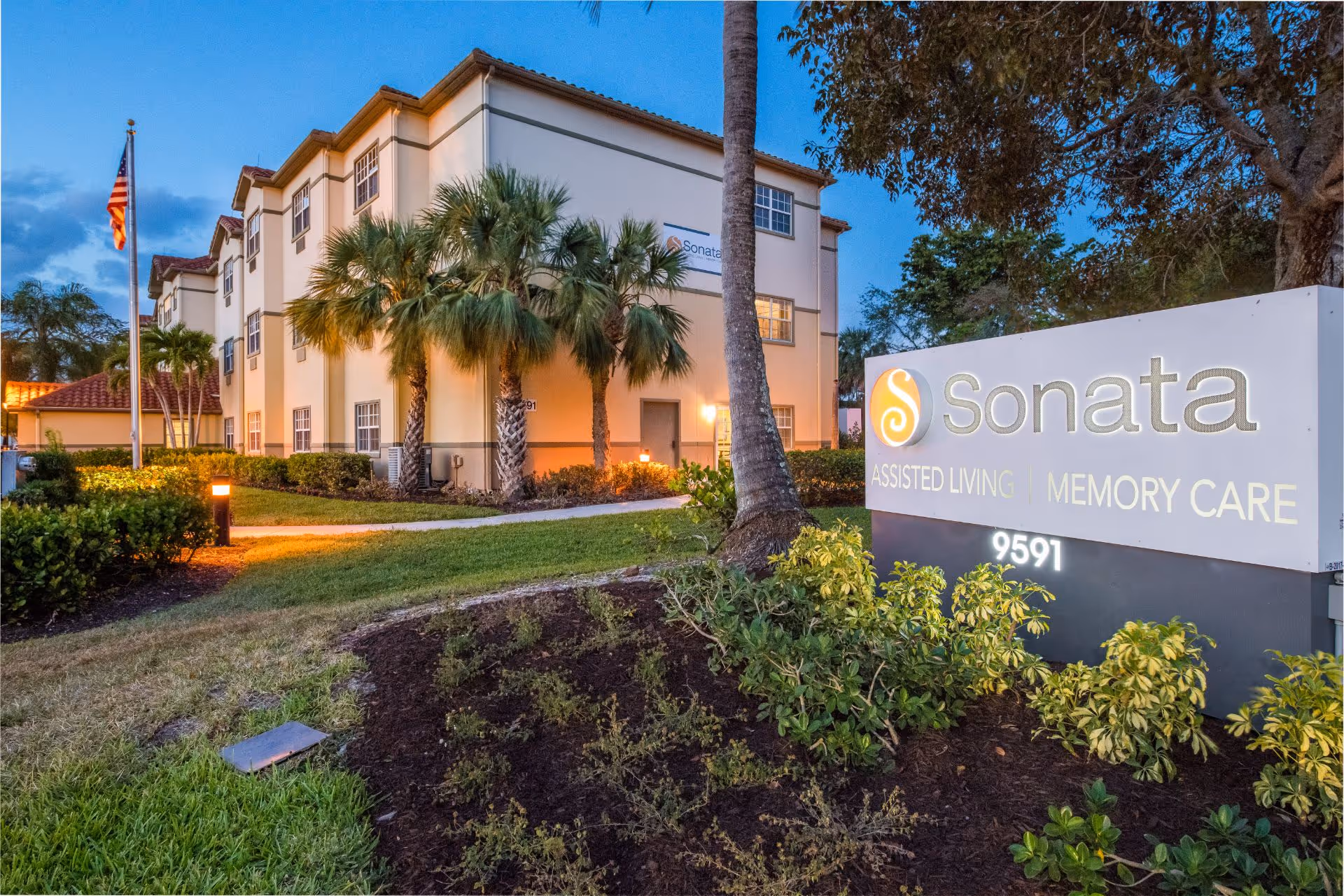 Exterior view of Sonata Boca Raton assisted living and memory care facility at dusk, showing a three-story building with palm trees, a well-maintained lawn, and a large illuminated sign with the facility name and address 9591.