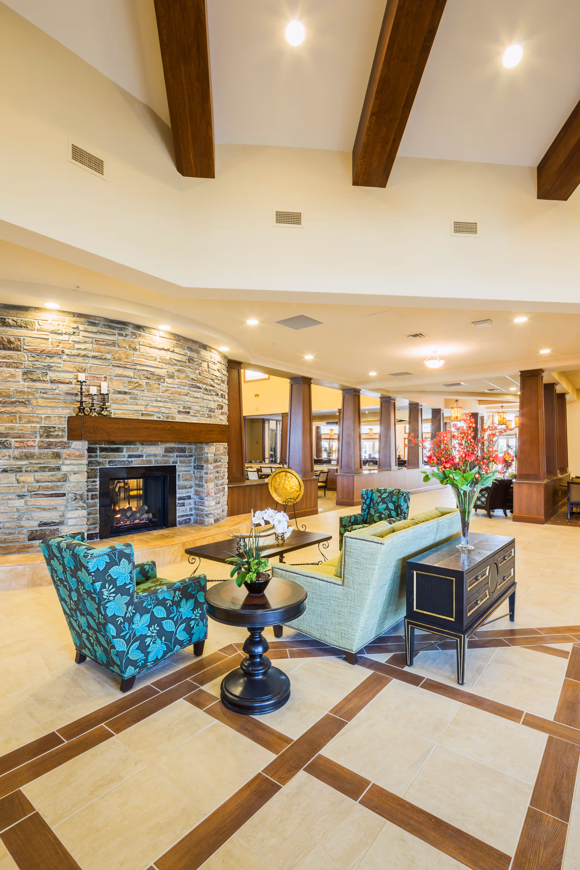 A spacious and well-lit senior living facility common area featuring a stone fireplace with a wooden mantel, two patterned armchairs, a light green sofa, a round side table with a plant, and a rectangular console table with a vase of red flowers. The room has a tiled floor with a wood inlay pattern and exposed wooden ceiling beams.