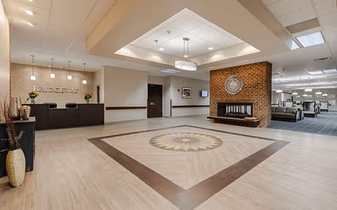 Spacious and well-lit lobby area of Bridgeway of Bensenville Supportive Living featuring a decorative floor design, a brick fireplace with a circular mirror above it, a reception desk with pendant lights, and comfortable seating in the background.