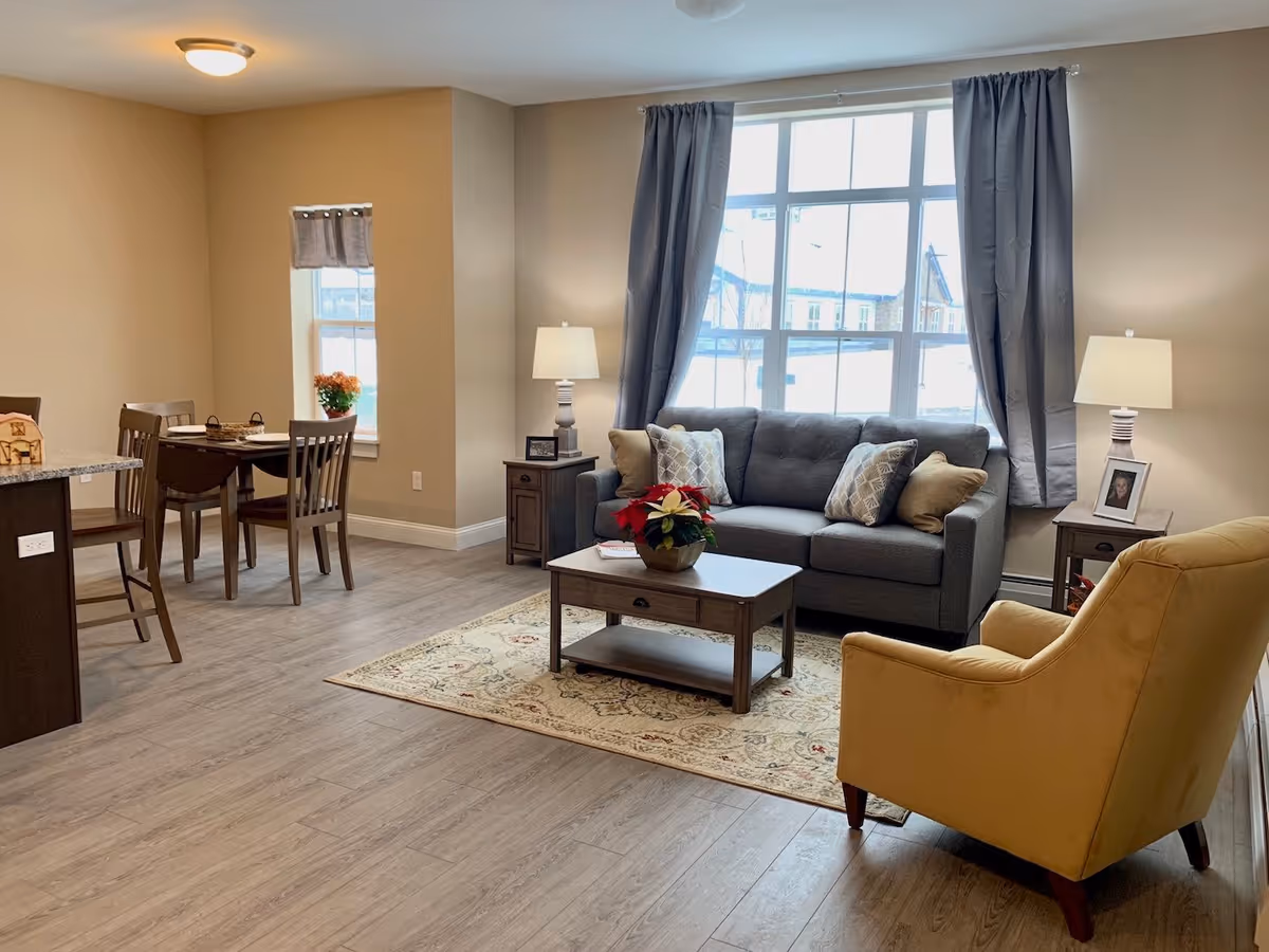 Open living area with a gray sofa, coffee table and yellow armchair by a large window, with a dining table visible in the background.