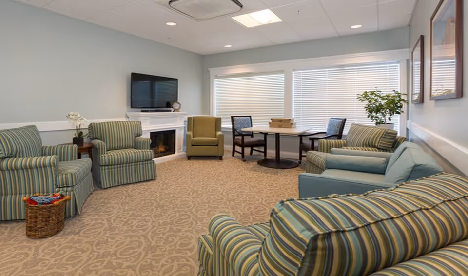 A cozy living room with striped armchairs and sofas arranged around a carpeted floor. There is a small round table with two chairs near a window with blinds. A flat-screen TV is mounted above a white fireplace, and there are plants and framed pictures on the walls.