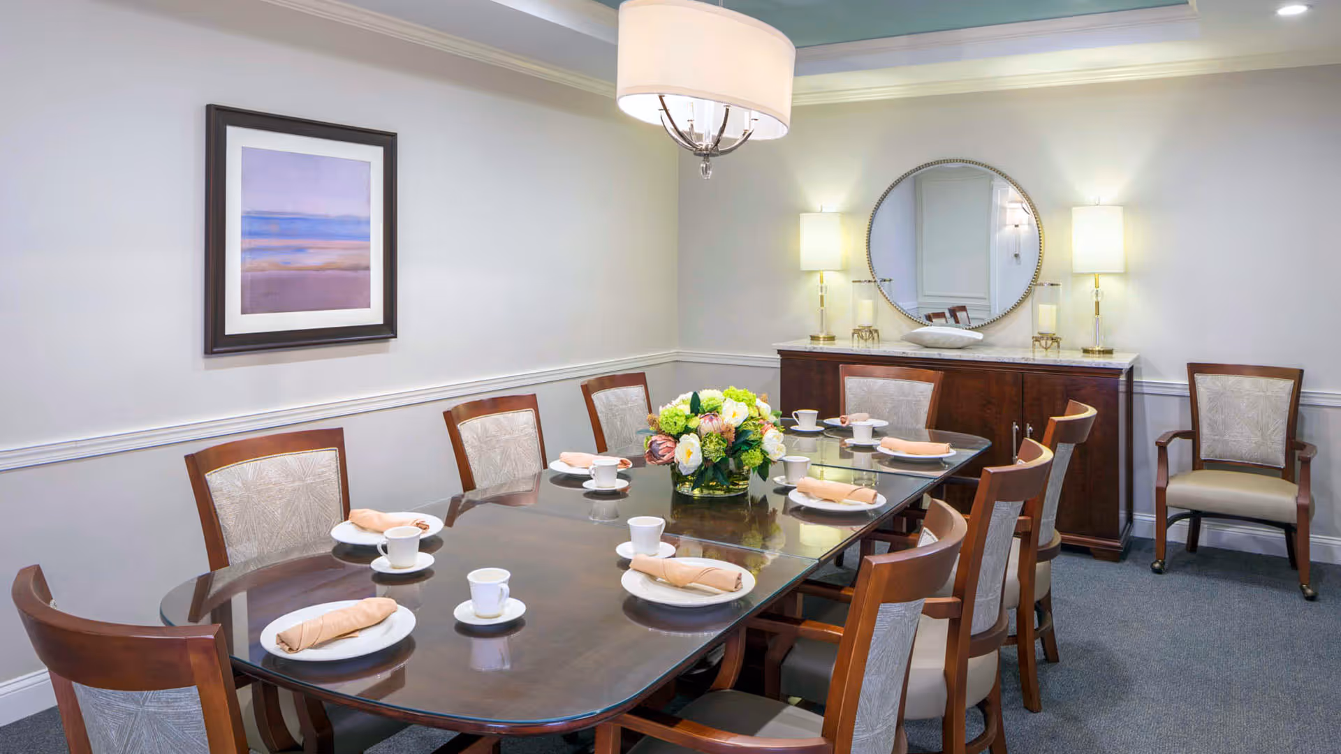 A formal dining room with a large wooden table set for eight people. Each place setting has a white plate, a cup and saucer, and a beige cloth napkin. A floral centerpiece is in the middle of the table. The room has light-colored walls, a framed landscape painting, a round mirror above a wooden sideboard with two lamps, and carpeted flooring.