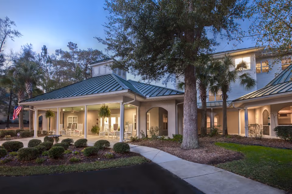 Exterior view of a senior living facility at dusk with a covered porch featuring rocking chairs and hanging plants. The building has a green metal roof, beige walls, and is surrounded by landscaped bushes, trees, and a walkway.