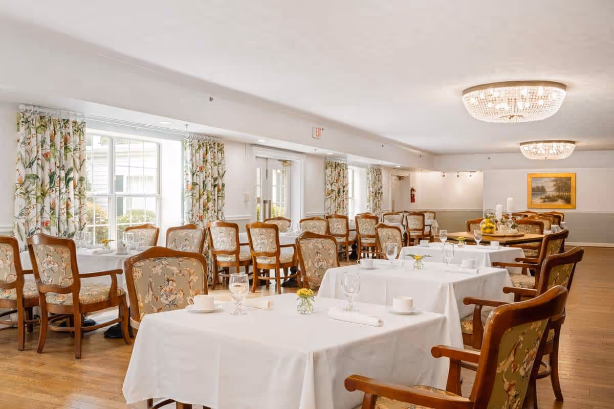 A bright and spacious dining room with multiple tables covered in white tablecloths, set with glasses, cups, and small flower vases. The room features wooden chairs with floral upholstery, large windows with floral curtains, and two elegant ceiling light fixtures. A painting hangs on the far wall.