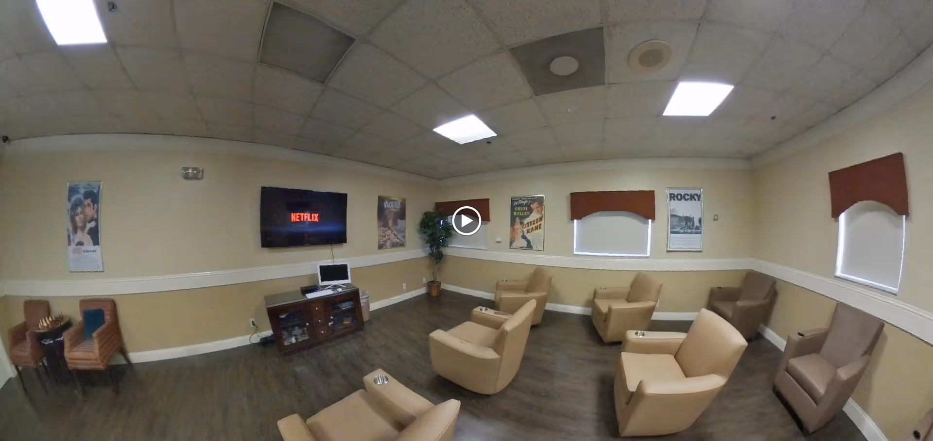 A cozy common room with several beige and brown armchairs arranged in a semi-circle facing a wall-mounted flat screen TV displaying the Netflix logo. The room has wood flooring, cream-colored walls with a white chair rail, and three windows with red valances. Movie posters are hung on the walls, and there is a small wooden cabinet under the TV with a computer on top. Two additional chairs and a small table are in the corner.