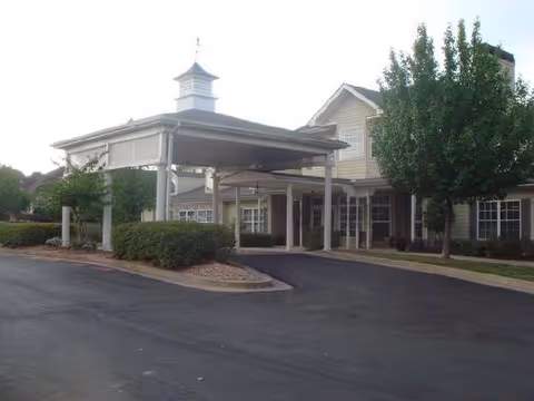Exterior view of a senior living facility named Sunabella of Towne Lake showing the front entrance with a covered drop-off area, surrounded by trees and bushes.
