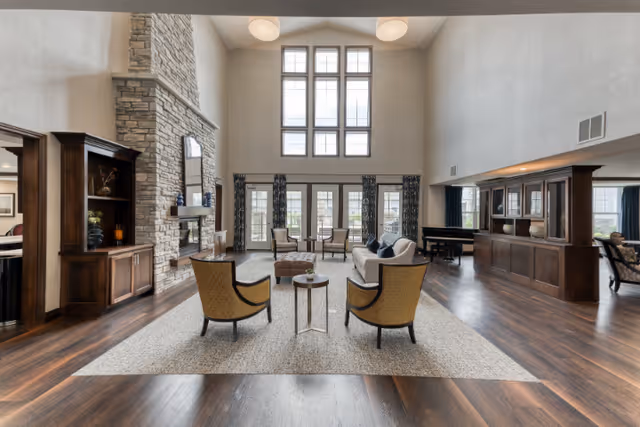 Spacious living room with high ceilings and large windows letting in natural light. The room features a stone fireplace, wooden built-in shelves, and a seating area with two armchairs, a sofa, and a coffee table on a large area rug. There is a piano and additional seating along the right side of the room.