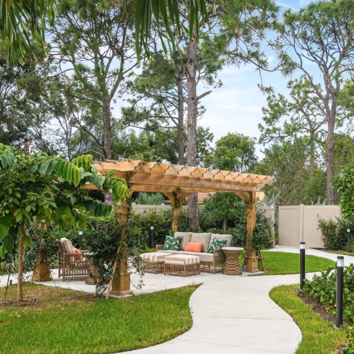 Outdoor garden area with a wooden pergola covering cushioned seating including a sofa, chairs, and ottomans. The space is surrounded by green grass, trees, and shrubs, with a curved concrete pathway leading to the seating area.