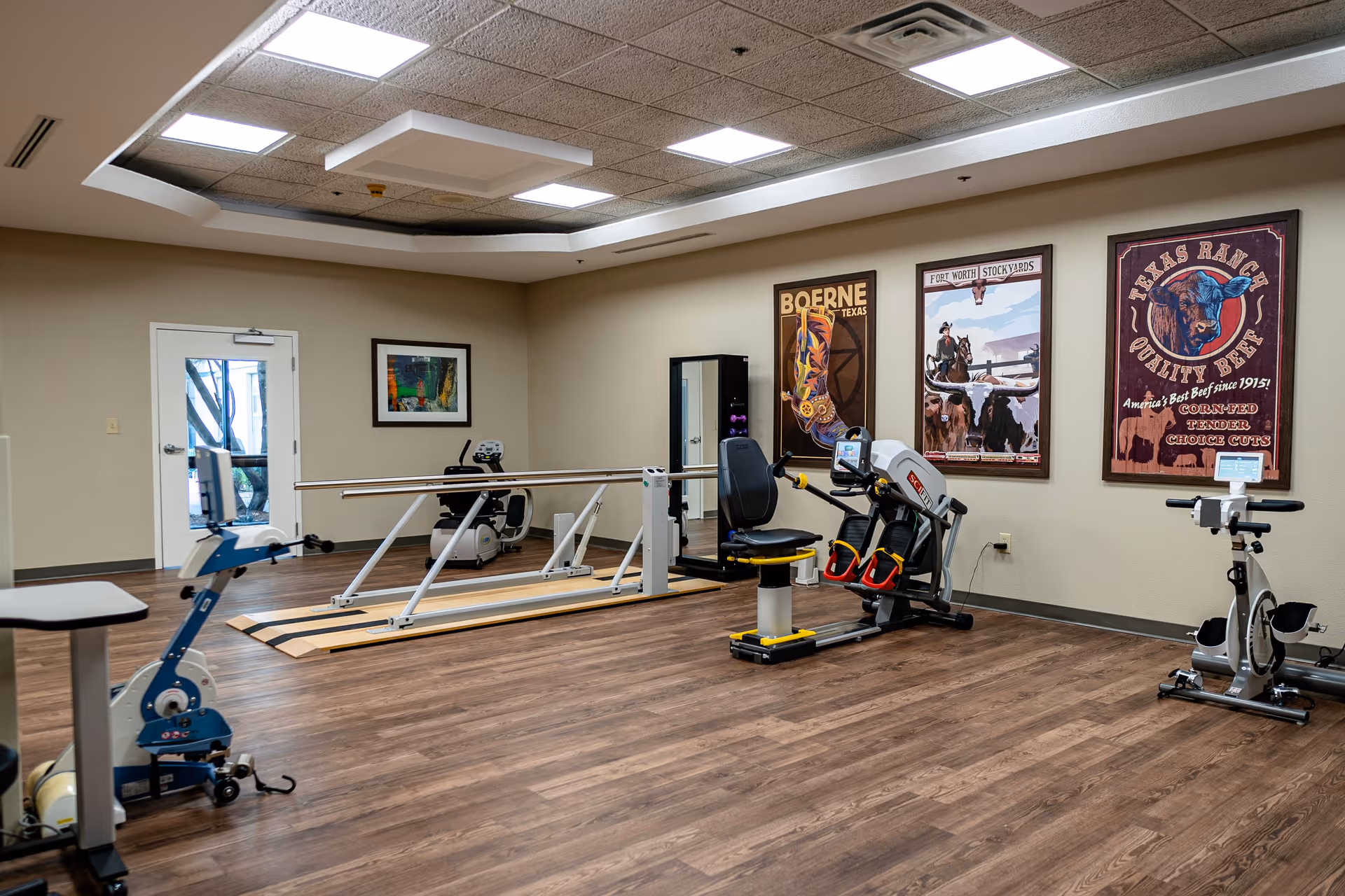 A rehabilitation gym room with parallel bars, exercise machines, and framed posters on the walls.