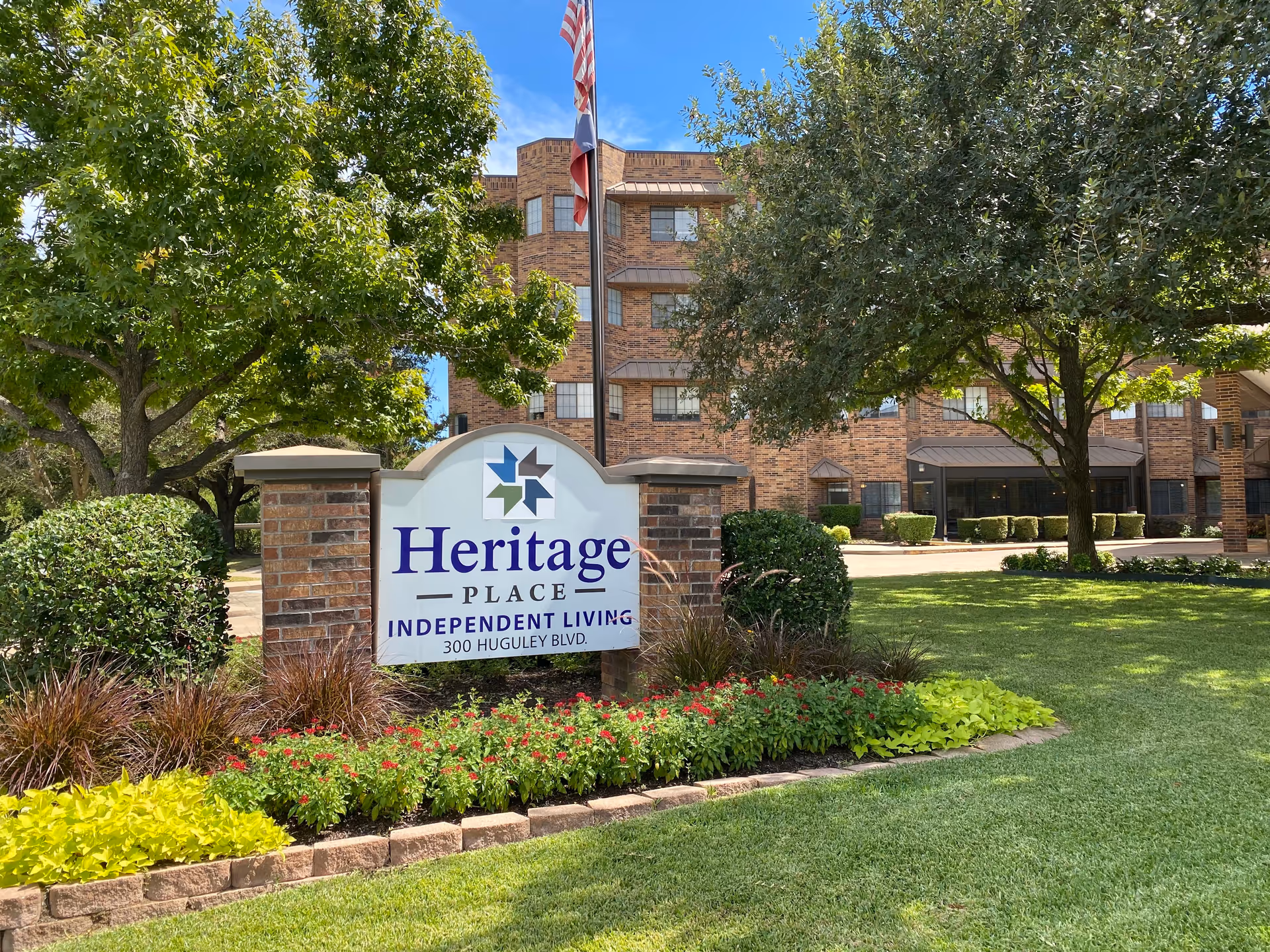 Sign reading 'Heritage Place Independent Living' in a landscaped front lawn with the brick senior living building behind it.