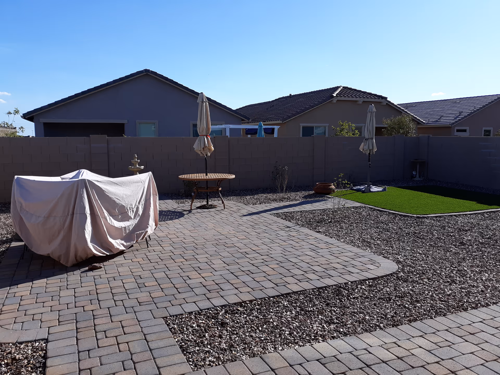 Backyard patio with a covered grill, round table and umbrellas on pavers, gravel landscaping and a small turf area in front of a block wall and neighboring houses.