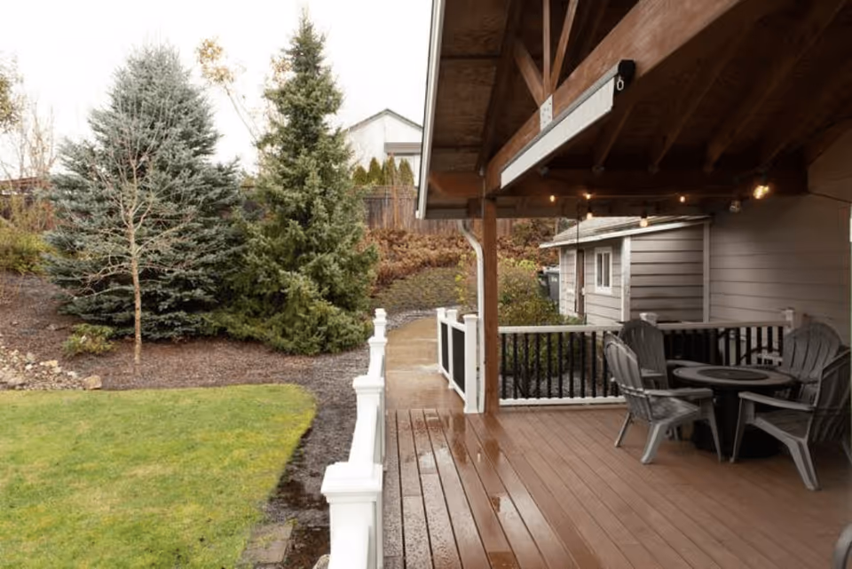 Covered wooden deck with a round table and chairs overlooking a grassy yard and evergreen trees.