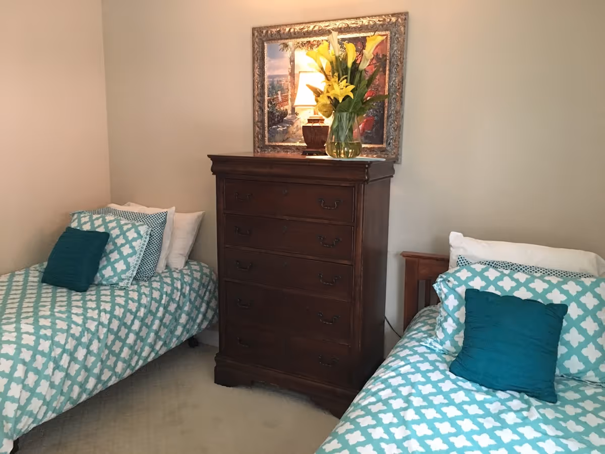 A bedroom with two twin beds featuring teal and white patterned bedding and teal accent pillows. Between the beds is a dark wooden chest of drawers with a vase of yellow flowers and a framed picture above it. The walls are light-colored and the floor is carpeted.